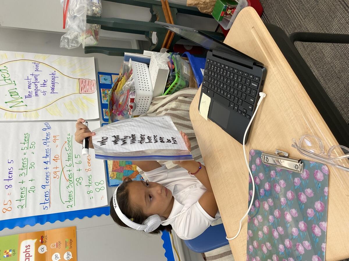 A student holding up a piece of paper encased in a Ziploc bag to show their tutoring on Zoom. They wrote the words: who, what, why where, there, here were, are, be, and have.
