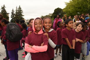 Two students pose and smile at the camera. A group of people watch near by.